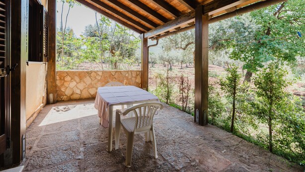 Een overdekt terras met een tafel en stoelen, stenen muren en uitzicht op bomen en landschap.