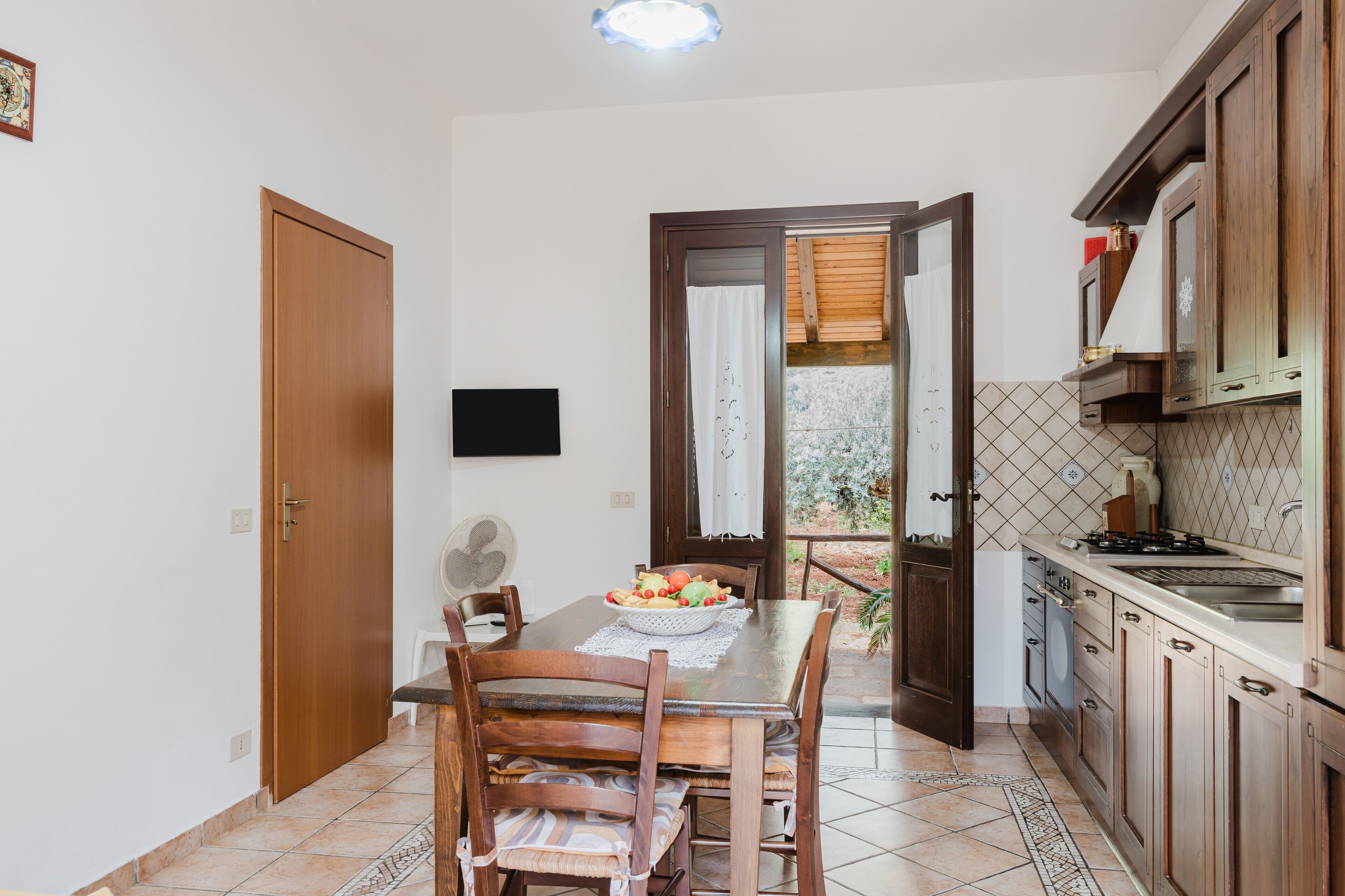 Une cuisine avec des meubles en bois, une table à manger avec des chaises, un téléviseur fixé au mur et, par une porte ouverte, une vue sur un balcon.