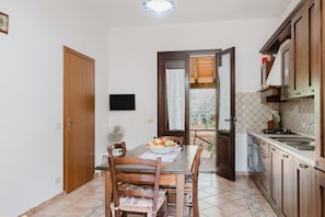 A kitchen with wooden cabinets, a dining table with chairs, a TV mounted on the wall, and a view through an open door to a balcony.