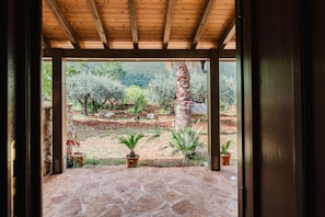 An open wooden structure with a stone floor, leading to a garden with potted plants and a view of a hilly landscape.