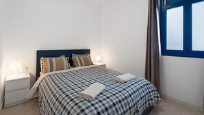 A neatly made bed with a checkered bedspread, two bedside lamps, a nightstand, and a window with curtains. - Apartment 'Playa Azul - 6' with Sea View, Private Terrace and Wi-Fi (Vallehermoso)