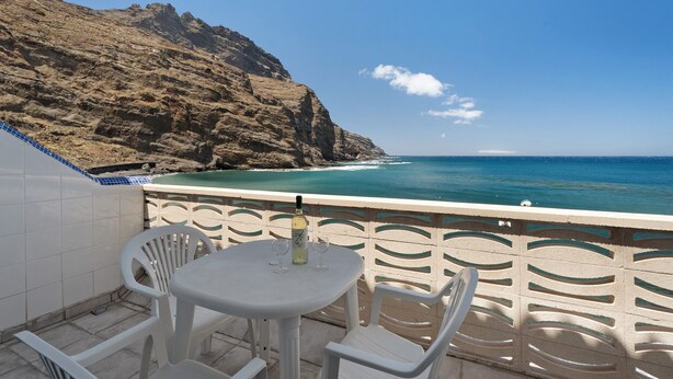 A balcony with a table and chairs, overlooking a coastal landscape with a rocky cliff and turquoise sea. - Apartment 'Playa Azul - 6' with Sea View, Private Terrace and Wi-Fi (Vallehermoso)