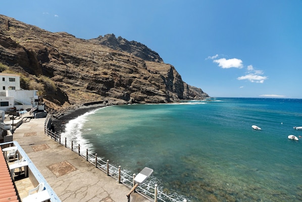 A coastal area with a rocky cliff, clear blue sea, and a building with outdoor seating.