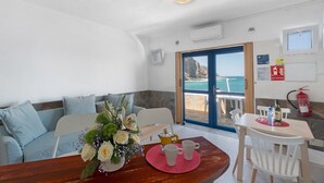 Een kamer met een bank, een tafel met een bloevas, een koffiehoek en uitzicht op de zee door een deur.