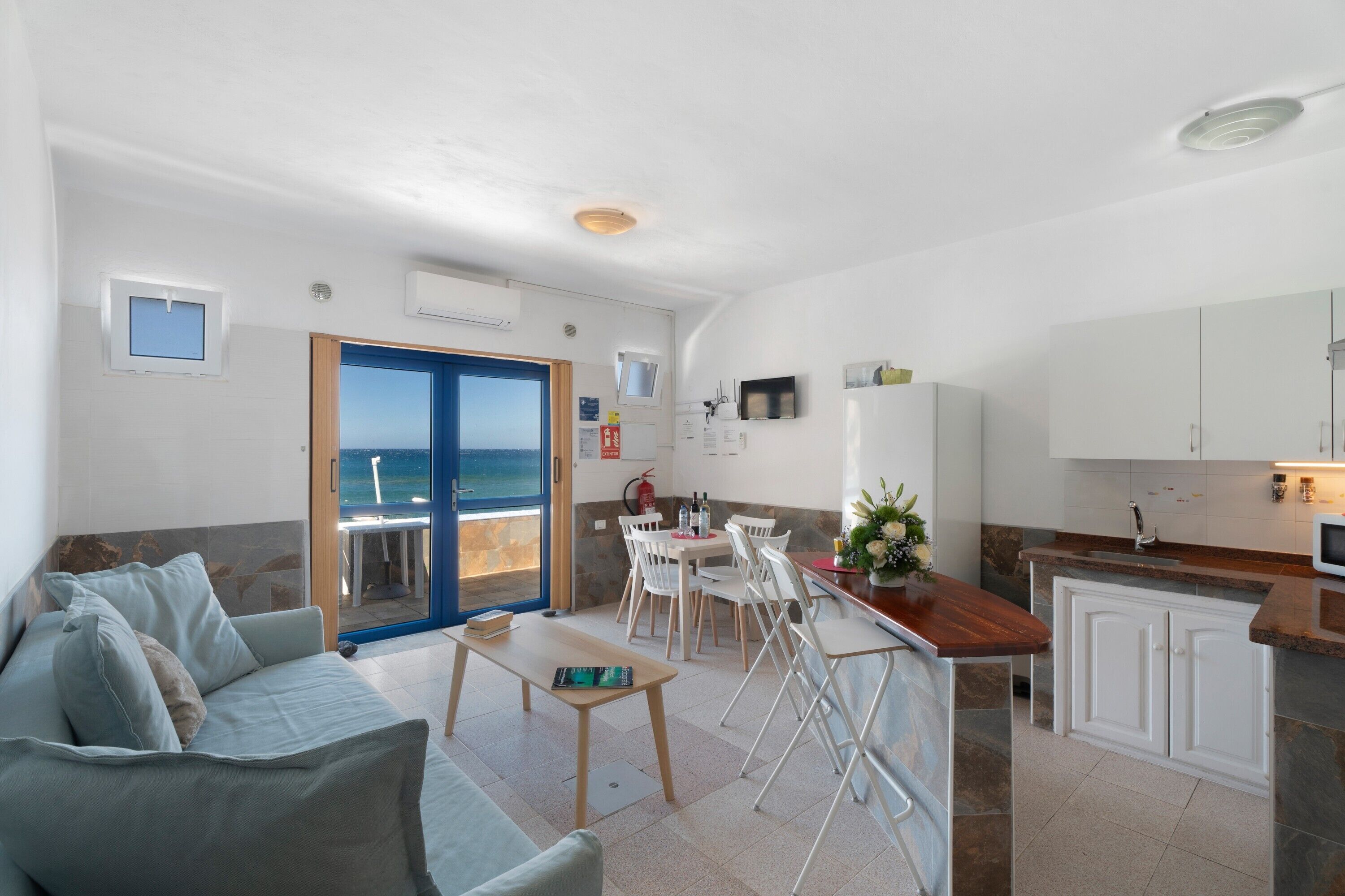 A modern living room with a sofa, a dining table, and chairs. There is a kitchen area with a counter and cabinets. A door leads to a balcony with a view of the sea.