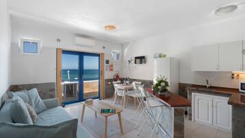 A modern living room with a sofa, a dining table, and chairs. There is a kitchen area with a counter and cabinets. A door leads to a balcony with a view of the sea.