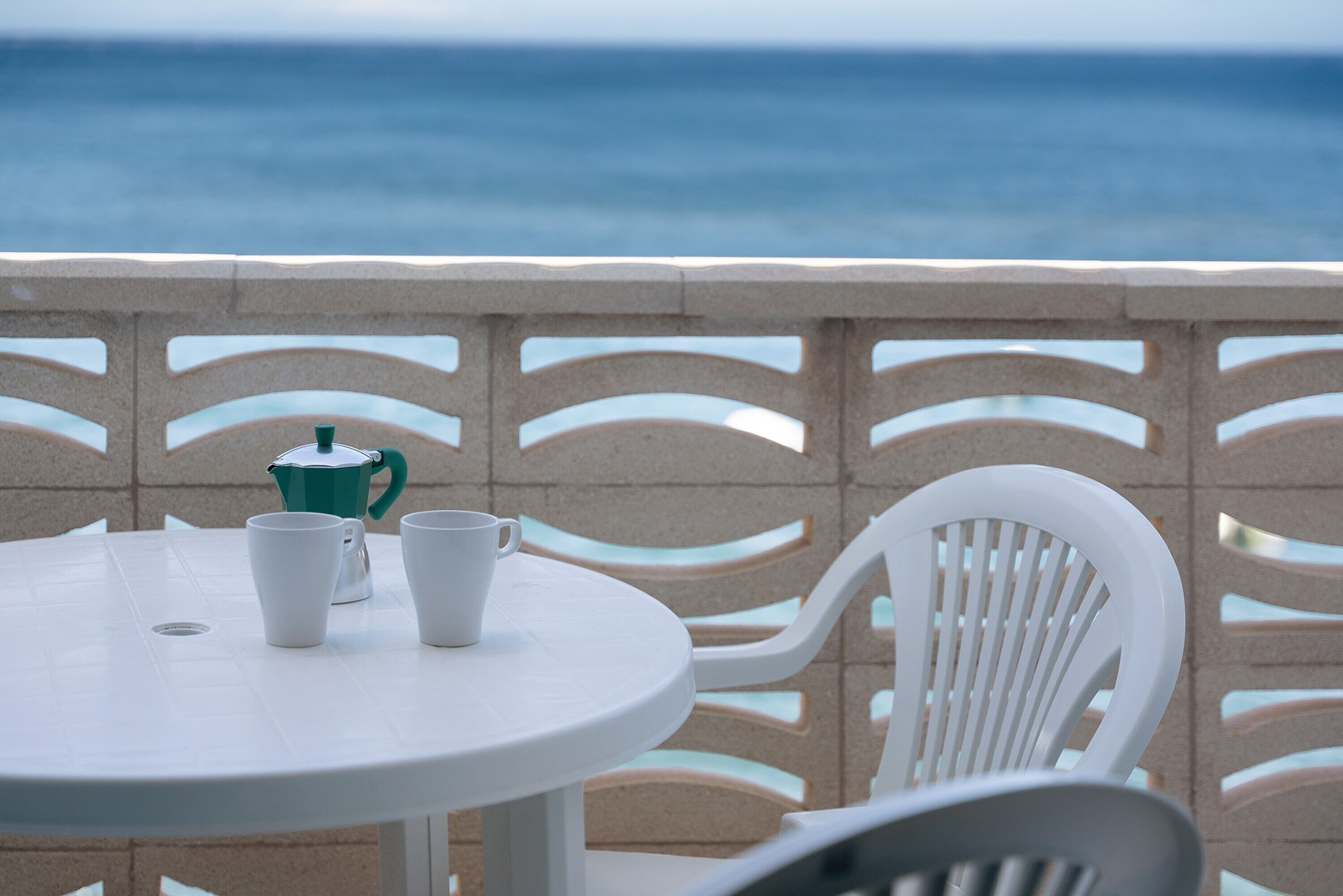 Une table ronde blanche, sur laquelle sont posées deux tasses blanches et un pot vert, le tout mis en valeur par un décor composé d’une vue sur la mer et d’une clôture à motifs.