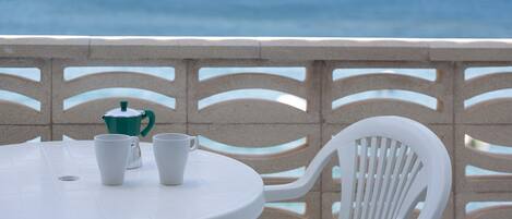 Une table ronde blanche, sur laquelle sont posées deux tasses blanches et un pot vert, le tout mis en valeur par un décor composé d’une vue sur la mer et d’une clôture à motifs.