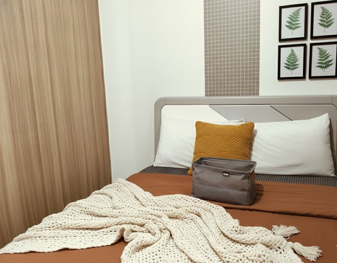 Un lit bien fait, avec une tête de lit grise, une couverture marron et un sac gris posé sur le lit.