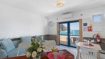 A room with a sofa, a table with a vase of flowers, a coffee setup, and a view of the sea through a door.