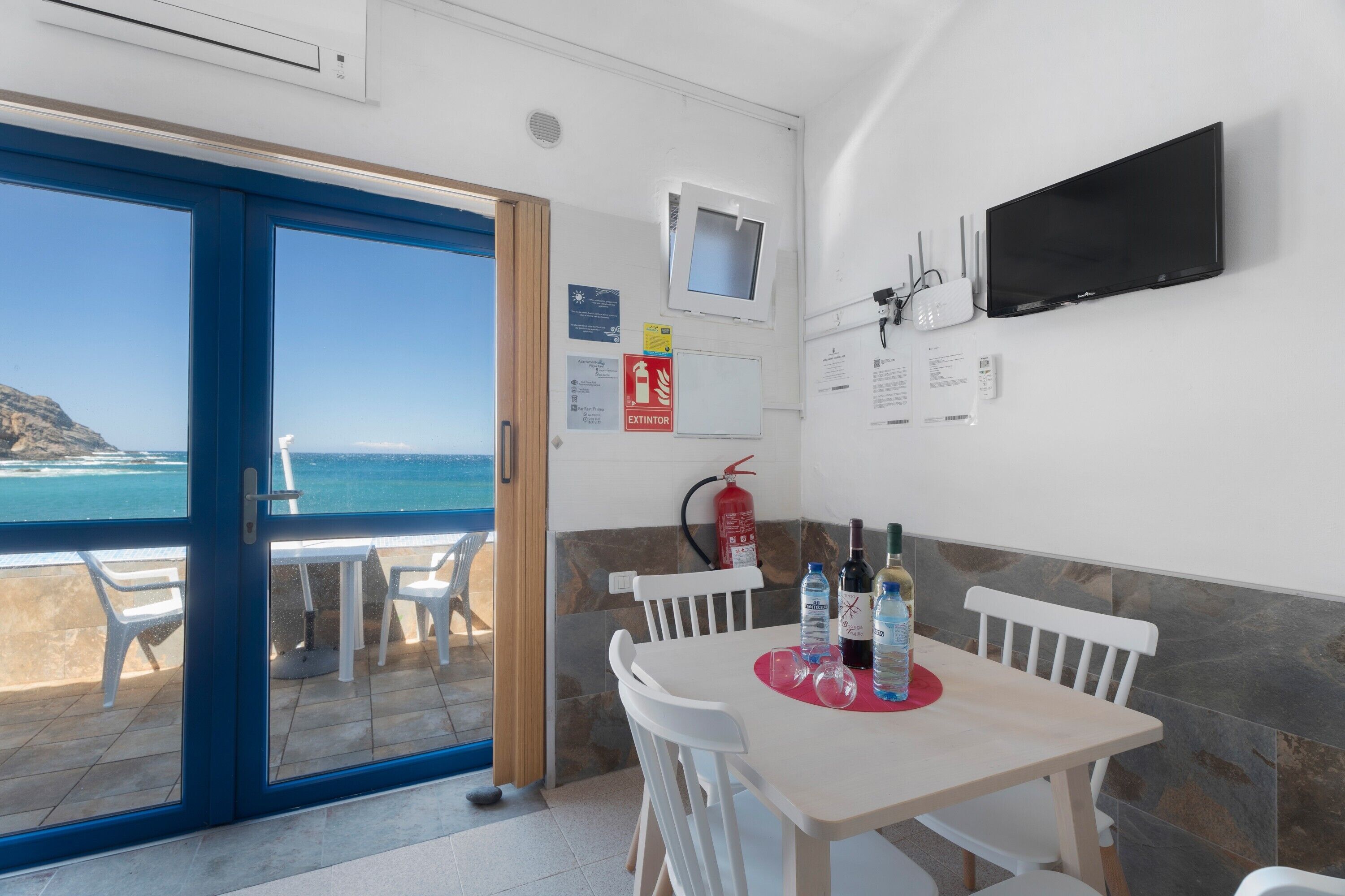 Una habitación con una mesa de comedor, sillas, un televisor de pantalla plana, un extintor y vistas al mar.