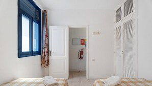 A room with two beds, a window with blue shutters, a door leading to a bathroom, and a fire extinguisher on the wall. - Apartment 'Playa Azul - 7' with Sea View, Private Terrace and Wi-Fi (Vallehermoso)