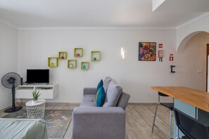 Una sala de estar moderna con un sofá gris, una mesita redonda, un televisor sobre un mueble blanco, un ventilador y una pared decorada con cuadros.