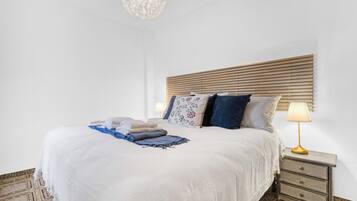 Un lit bien fait, avec une couette blanche, un plaid bleu et plusieurs oreillers. Il y a une table de chevet avec une lampe et un tiroir. La chambre est dotée d’une tête de lit en bois et d’un tapis à motifs sur le sol.