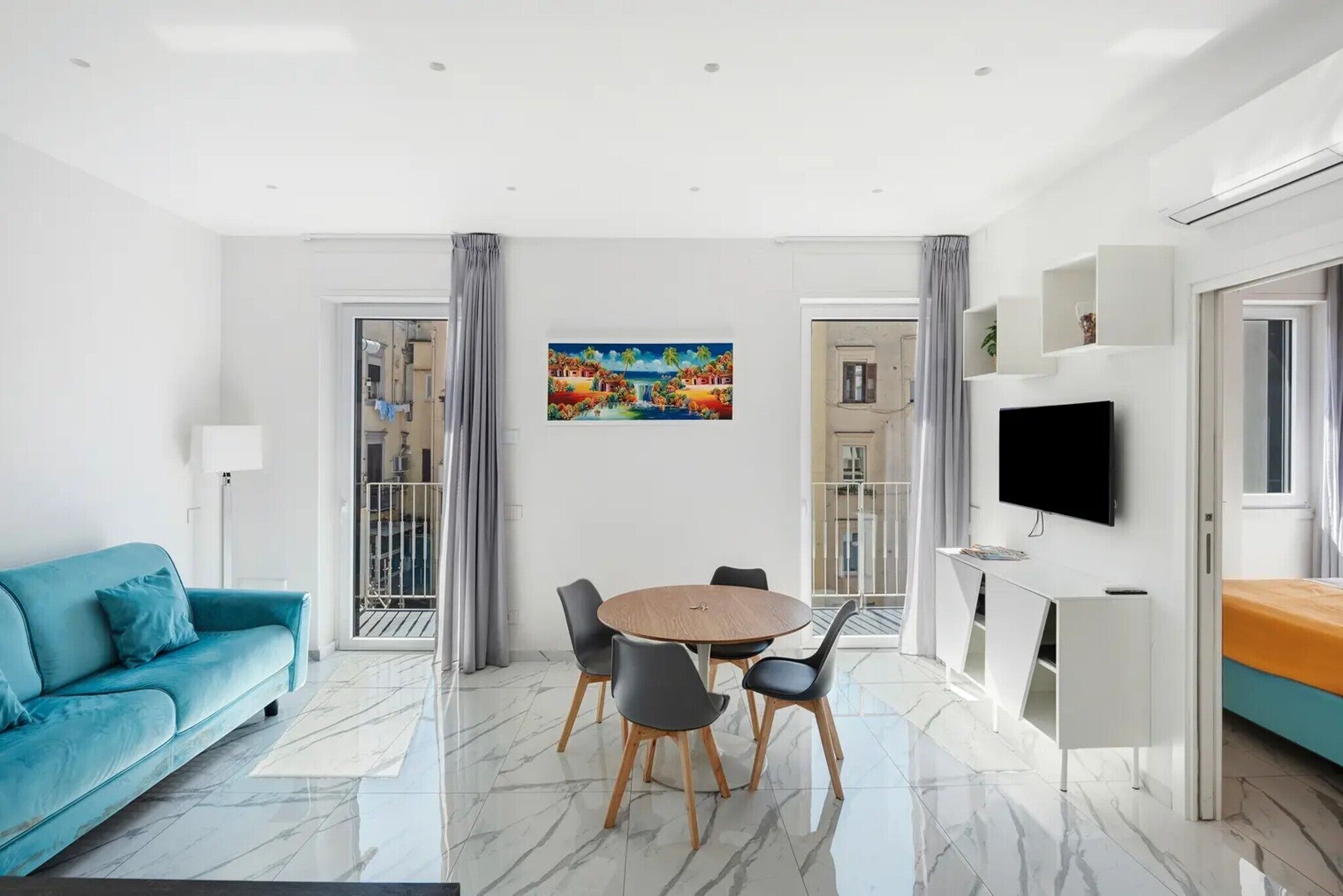 A modern living room with a blue sofa, a round dining table with chairs, a TV mounted on the wall, and a painting above the table.