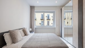 Ein Schlafzimmer mit einem großen Bett, einem Nachttisch, einem Fenster mit Blick auf Gebäude und einer Schiebetür zum Balkon.