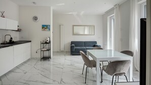 Eine moderne Küche mit weißen Schränken, einem Marmorboden, einem Essbereich mit Glastisch und Stühlen, einem blauen Sofa und einem großen Spiegel.
