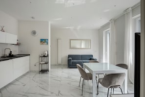 A modern kitchen with white cabinets, a marble floor, a dining area with a glass table and chairs, a blue sofa, and a large mirror.