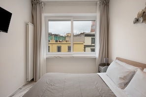 Un dormitorio con cama, ventana con cortinas, mesita de noche con lámpara y televisor montado en la pared.