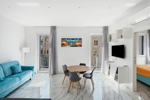 A modern living room with a blue sofa, a round dining table with chairs, a TV mounted on the wall, and a painting above the table.