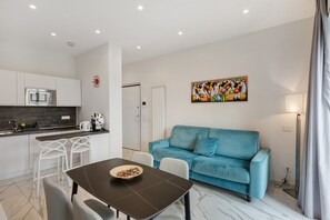 A modern kitchen with a microwave, white bar stools, a dining table with a bowl, a blue sofa, and a painting on the wall.