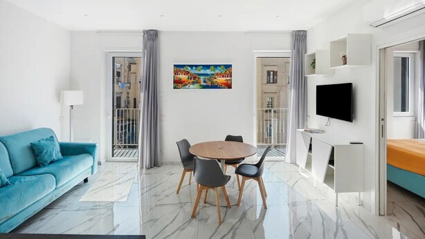 Una sala de estar moderna con un sofá azul, una mesa de comedor redonda con sillas, un televisor montado en la pared y un cuadro sobre la mesa.
