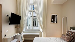 Quarto com cama, escrivaninha, cadeira, televisão e janela com cortinas.