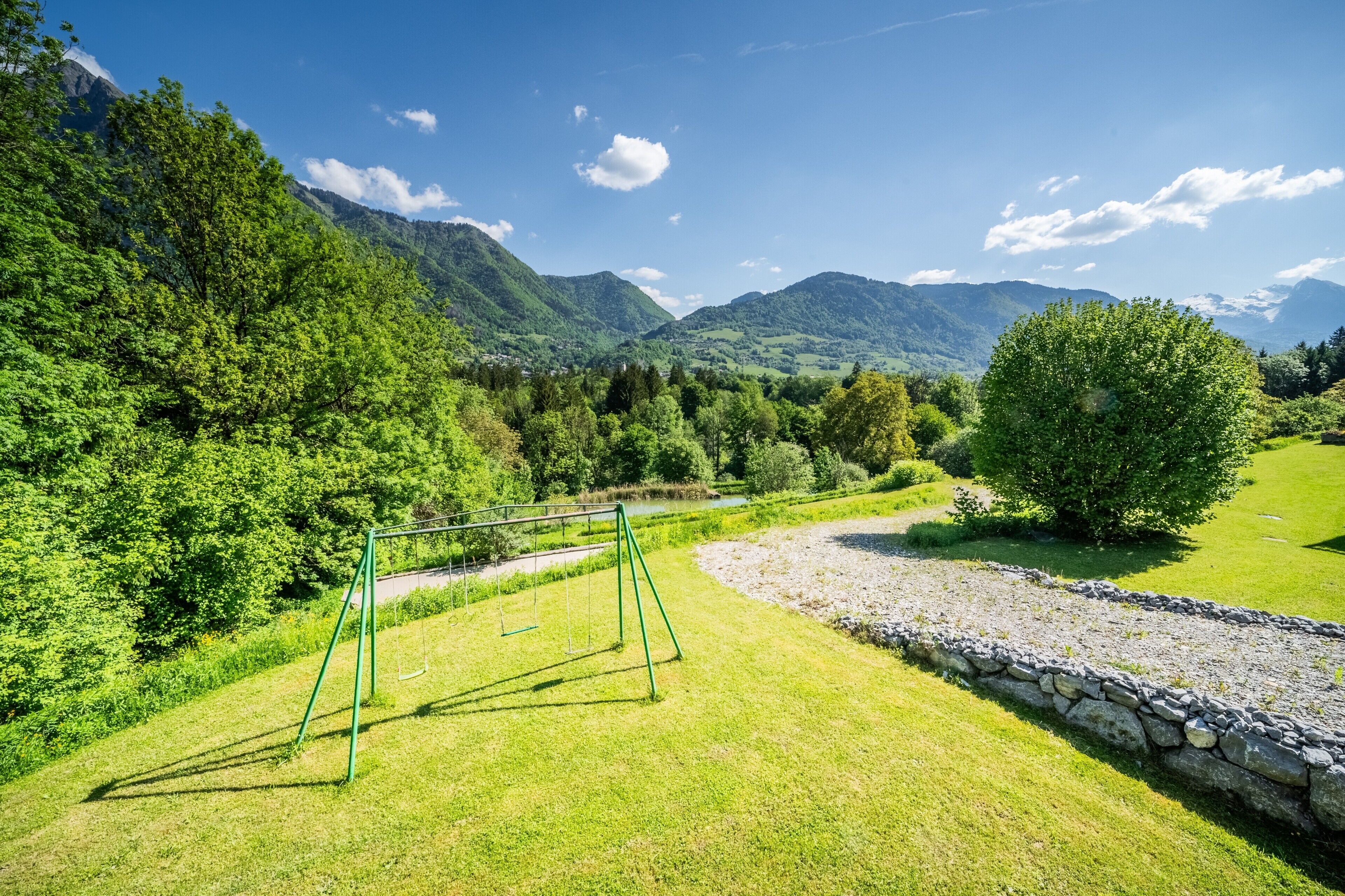 Un'area verde con altalene, un muro in pietra e montagne sullo sfondo.