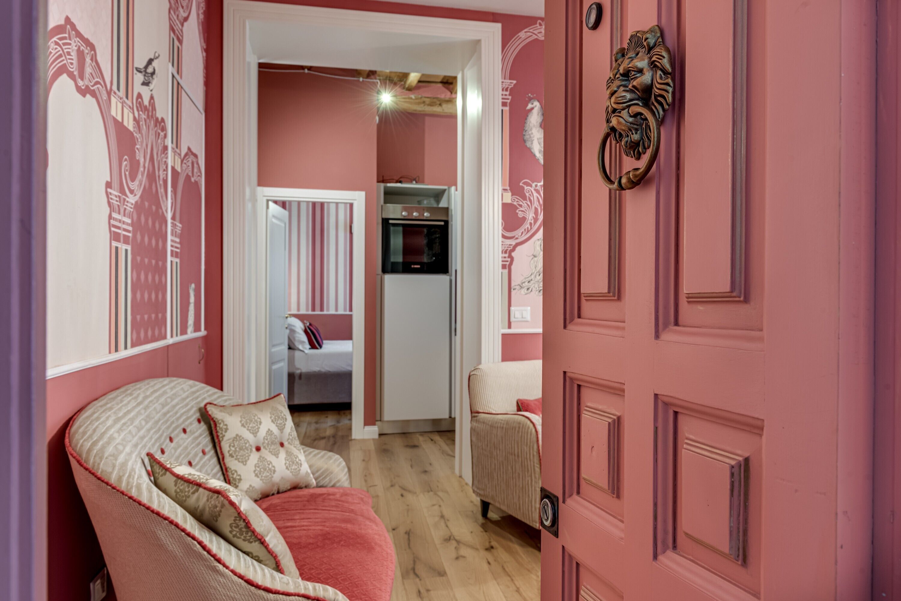 A pink-themed room with a bed, a chair, a door with a lion head knocker, and a kitchen area with a refrigerator and oven.