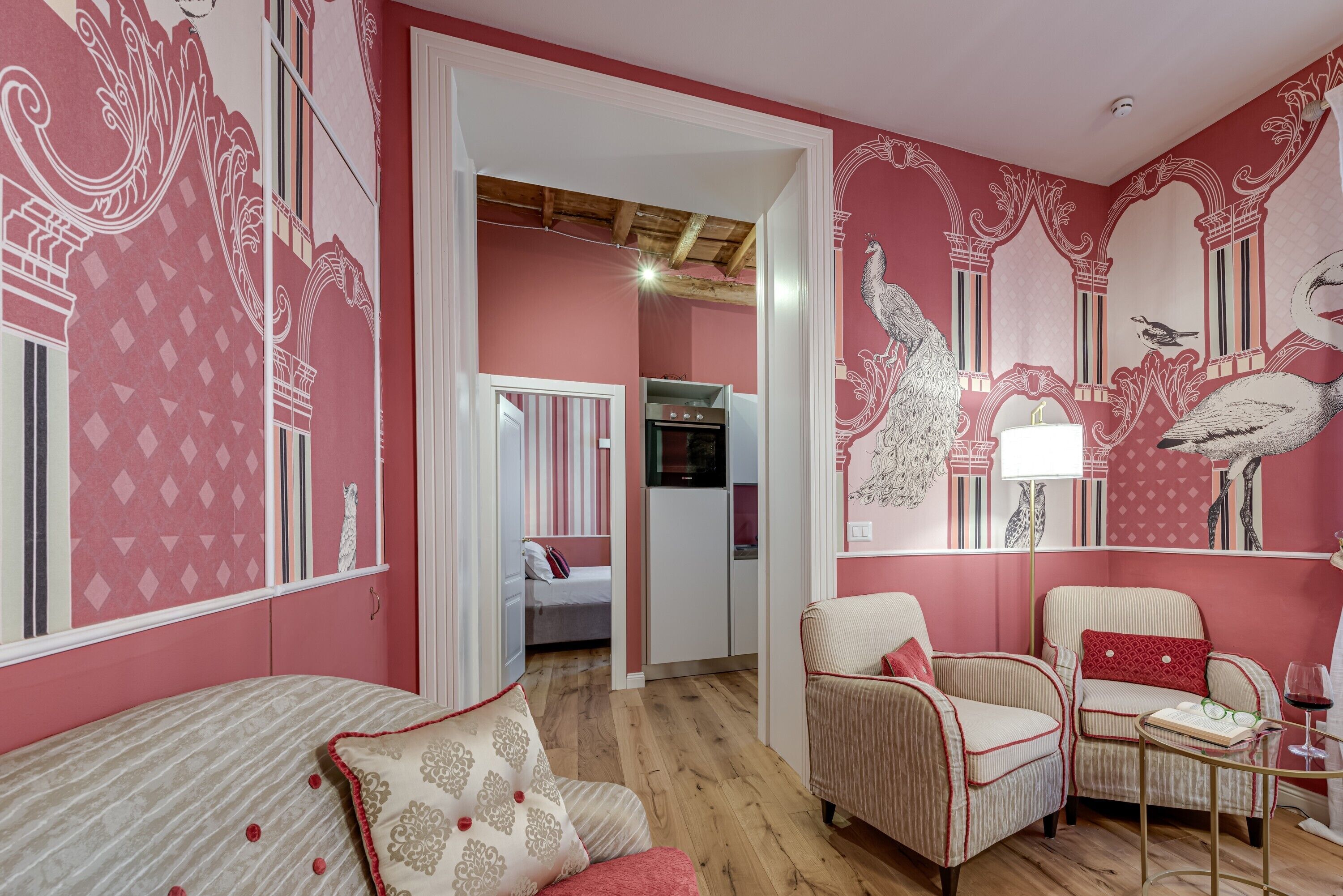 A room with a bed, two chairs, a small table, and a mirror. There is a door leading to another room.