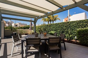 Eine Terrassesituation im Freien mit Tisch und Stühlen, einem Pergola darüber und einer Hecke im Hintergrund.