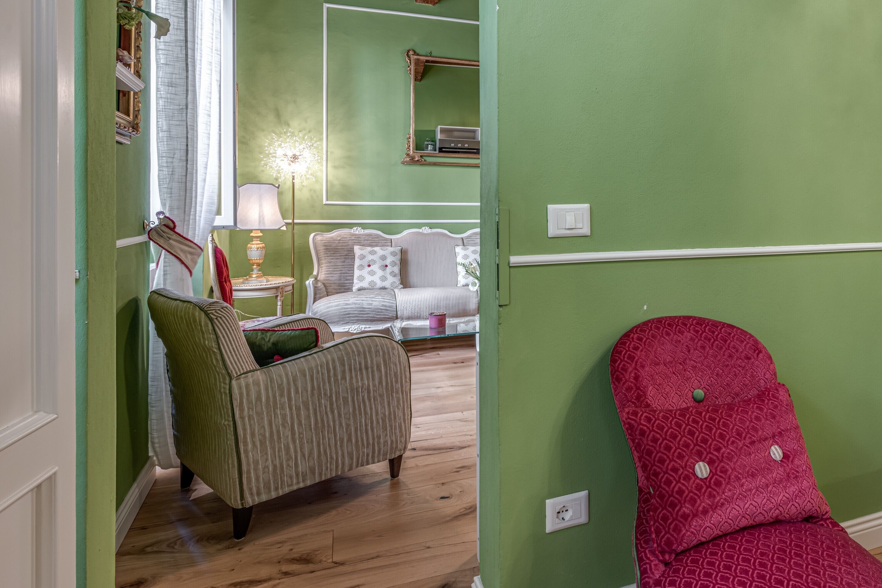 A room with a green wall, a wooden floor, a striped armchair, a white sofa, and a red chair.