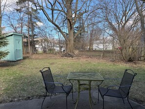 Une terrasse avec deux chaises en métal et une table, un petit abri de jardin et un grand arbre dans un jardin.
