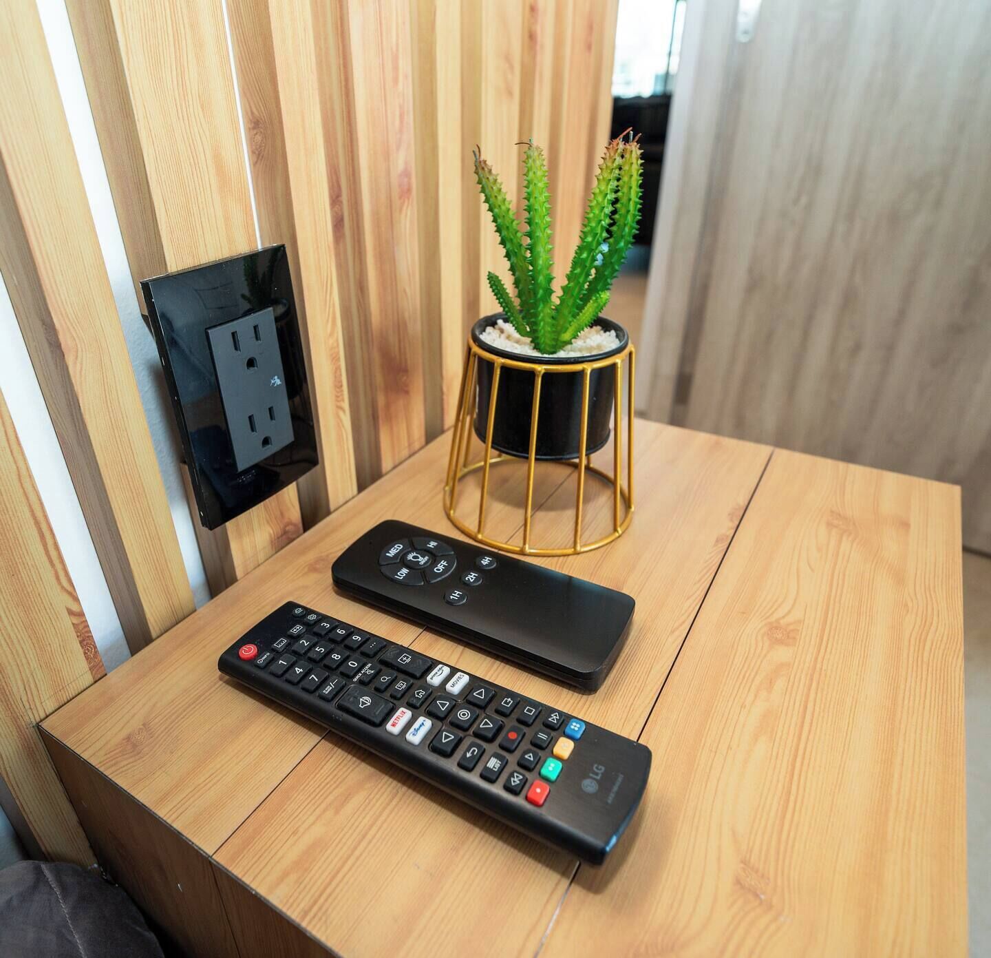 A wooden table with a remote control, a power outlet, and a potted plant.