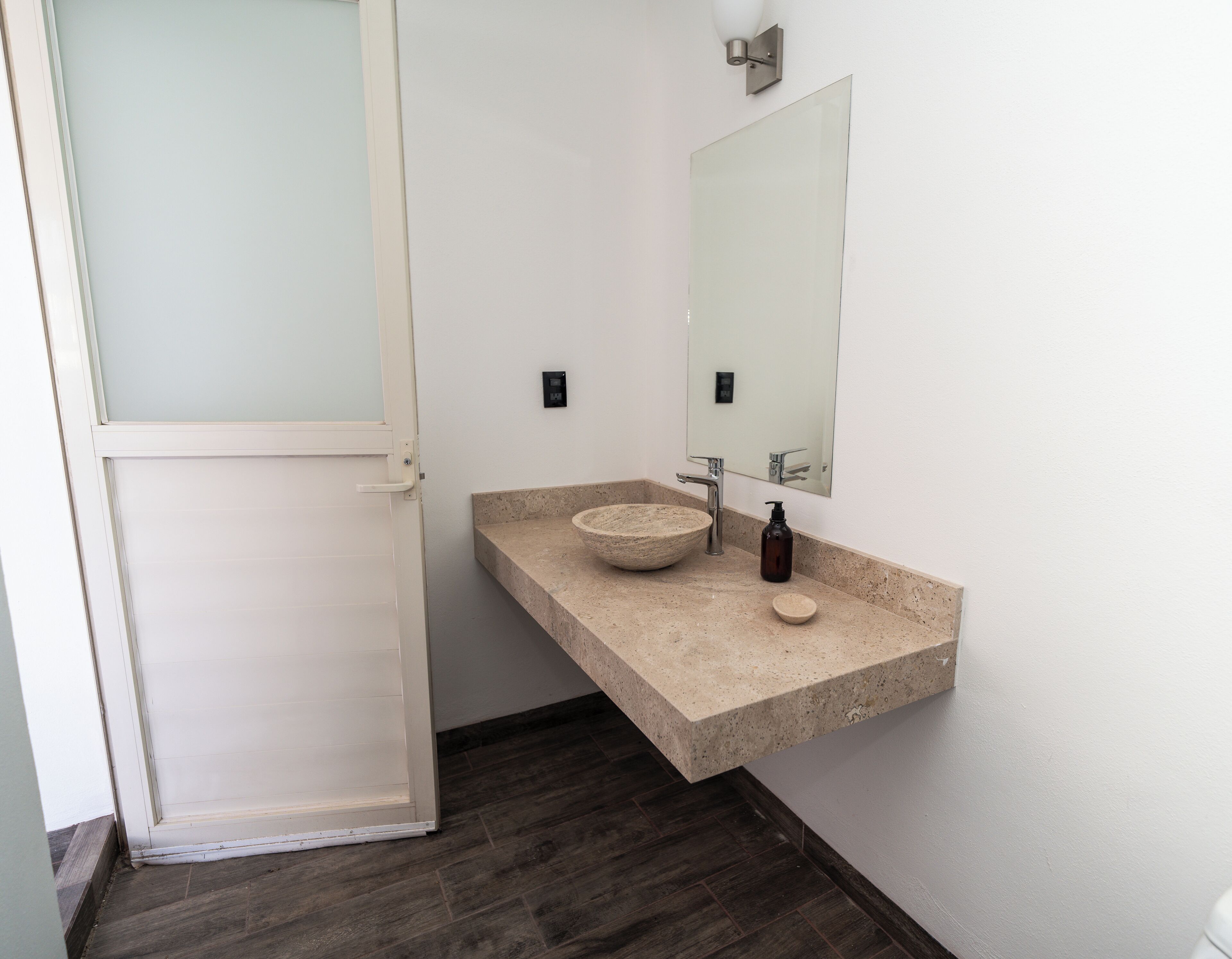 A modern bathroom with a white door, a beige countertop, a large bowl sink, a mirror, and a dark wooden floor.