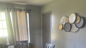 Un comedor con una mesa de madera, sillas negras y una planta en maceta. Hay cortinas en la ventana, y se ve una puerta al fondo.