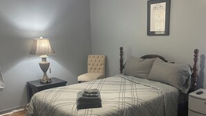 Un dormitorio con cama, una silla, una mesita de noche, una lámpara y un cuadro en la pared.