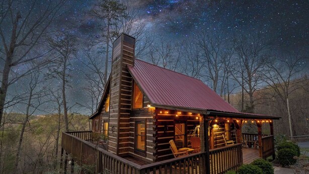 A log cabin with a red roof, a deck with outdoor seating, and a view of the night sky. - Charming Cabin in the Smokies -close to attractions, Hot Tub, Pool Table, Arcade (Sevierville)