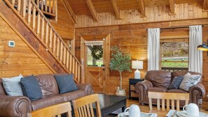 A wooden cabin interior with a staircase, two leather sofas, a dining table set with plates and cups, and a view of the outdoors through a window. - Charming Cabin in the Smokies -close to attractions, Hot Tub, Pool Table, Arcade (Sevierville)