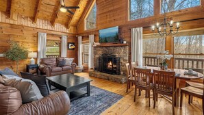 A cozy cabin interior with a stone fireplace, wooden furniture, and large windows. - Charming Cabin in the Smokies -close to attractions, Hot Tub, Pool Table, Arcade (Sevierville)