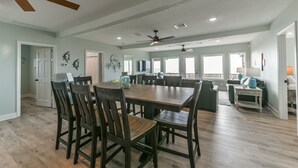 A spacious dining area with a large wooden table and chairs, a ceiling fan, and a view of the ocean through large windows.