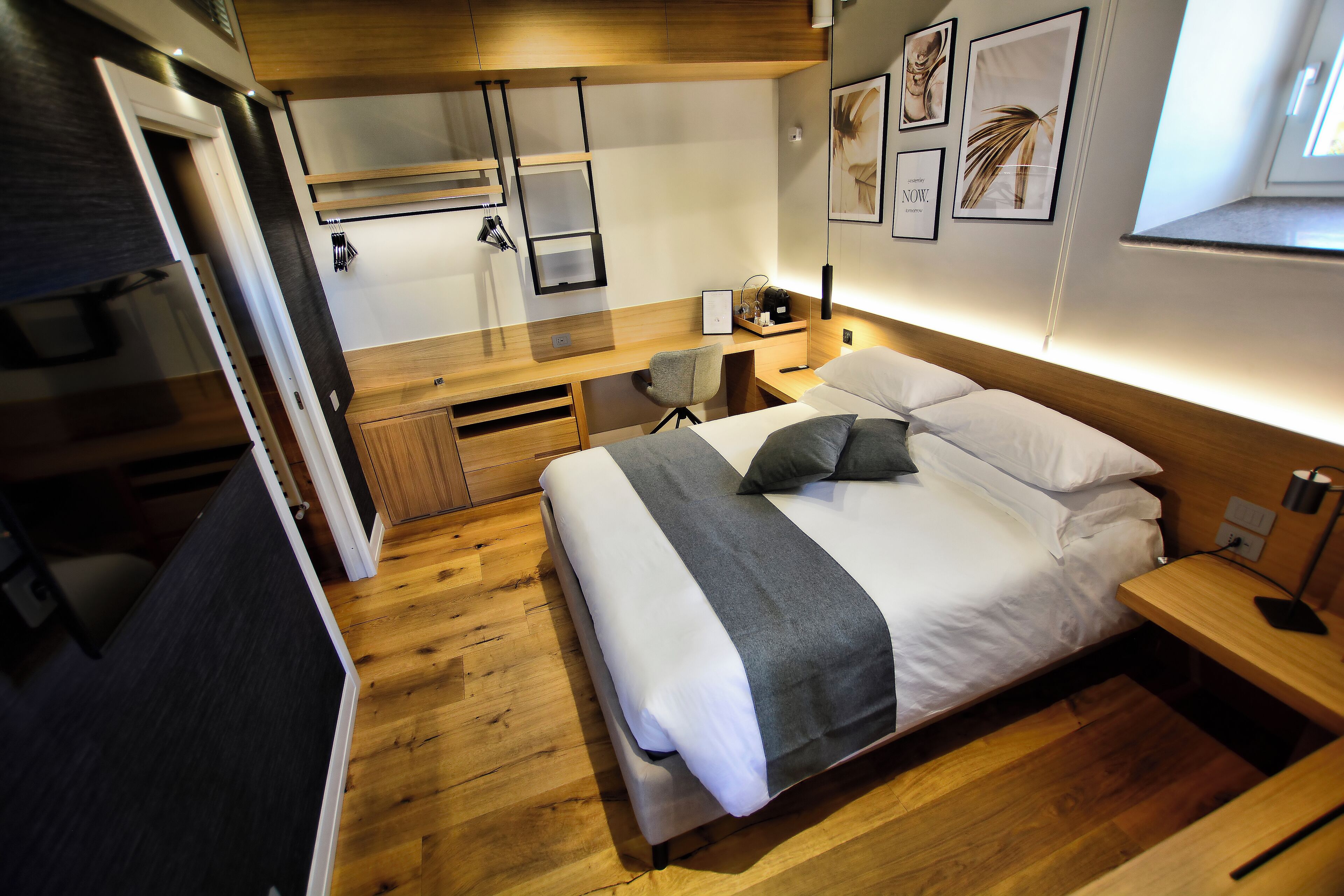 Una habitación de hotel moderna con piso de madera, una cama con ropa blanca, un escritorio con silla y obras de arte en las paredes.