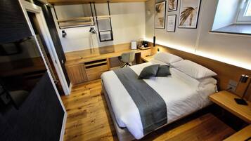 Una habitación de hotel moderna con piso de madera, una cama con ropa blanca, un escritorio con silla y obras de arte en las paredes.