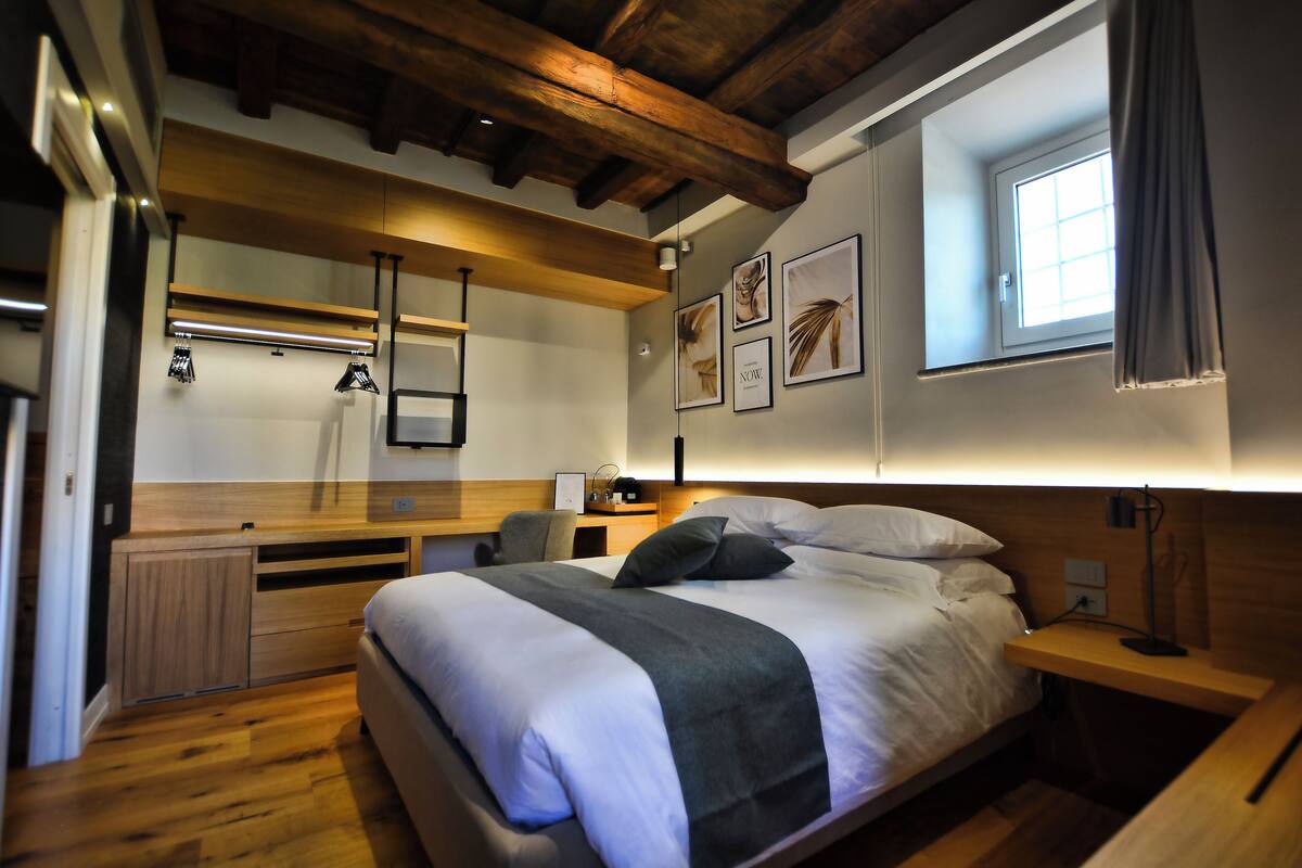Un dormitorio moderno con una cama grande, vigas de madera en el techo, una mesita de noche y un ventanal con cortinas.