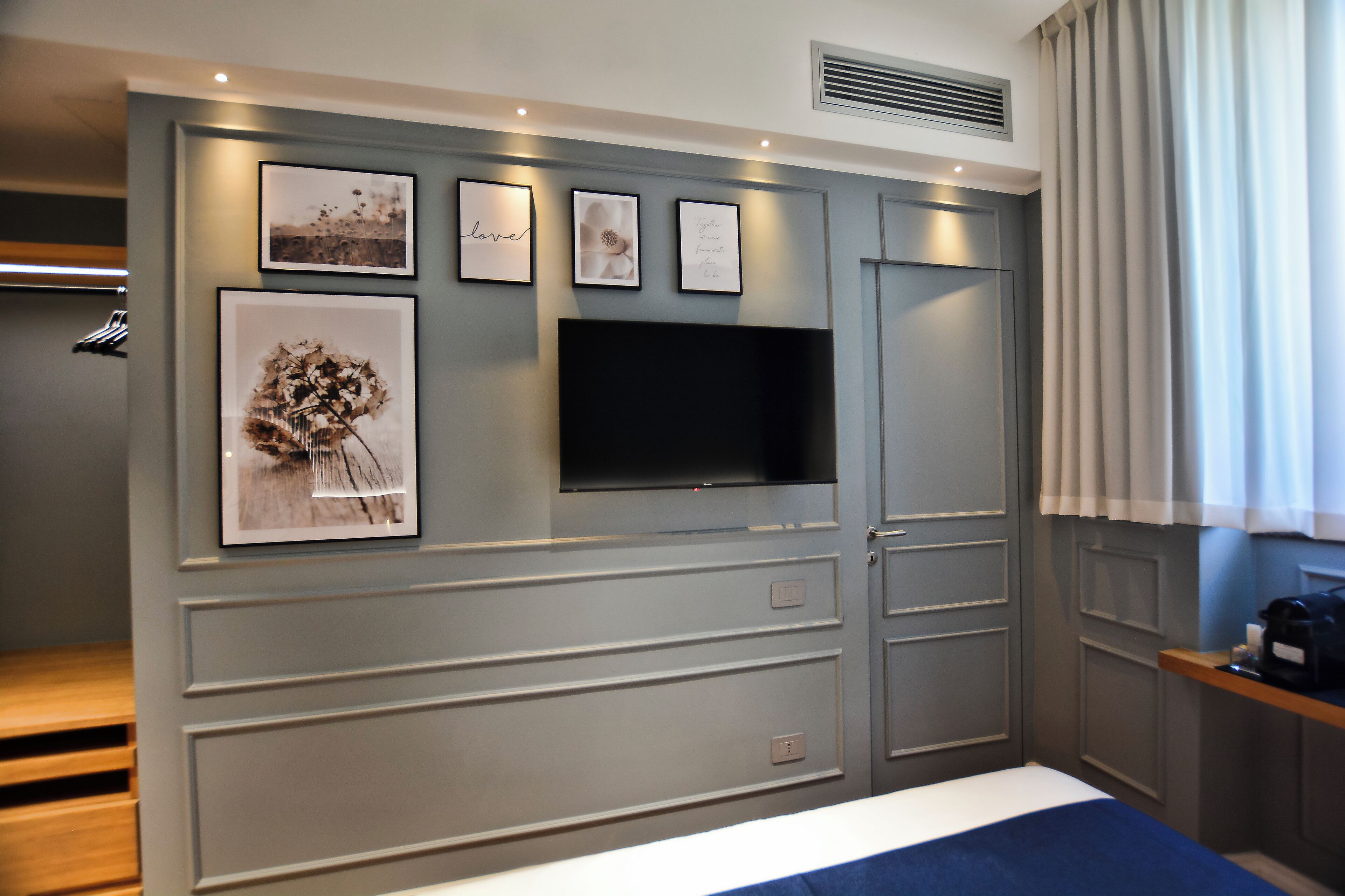 A modern bedroom with a grey wall, a flat-screen TV, and framed artwork.