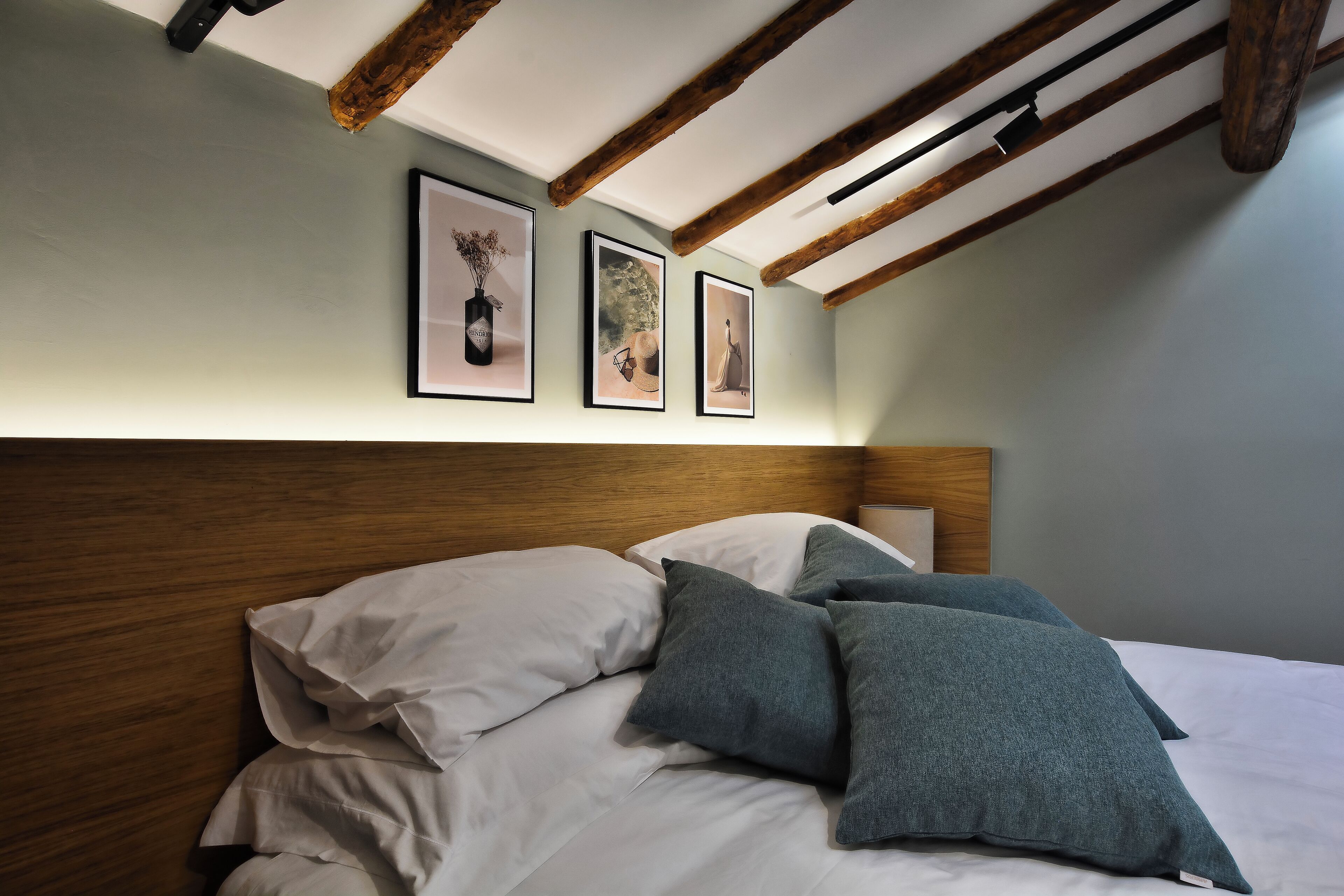 Un dormitorio con cabecera de madera, cama con ropa de cama blanca y gris, y tres cuadros enmarcados en la pared.