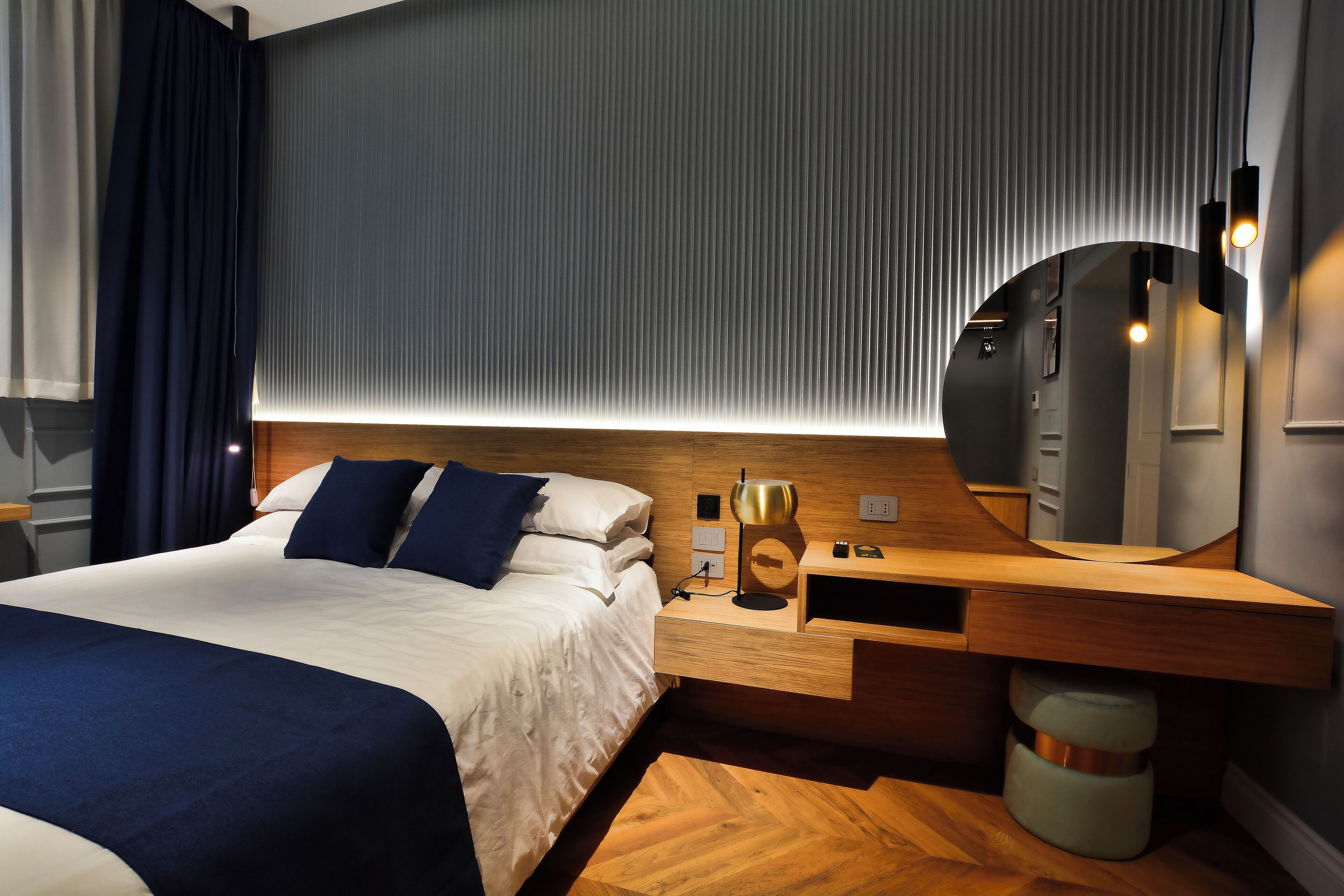 Moderni hotellihuone, jossa on suuri sänky, puinen yöpöytä, peili ja seinään kiinnitetty valaisin.