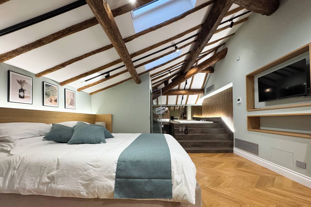 Un dormitorio con una cama grande, vigas de madera, un televisor montado en la pared y una escalera que conduce a una zona superior.