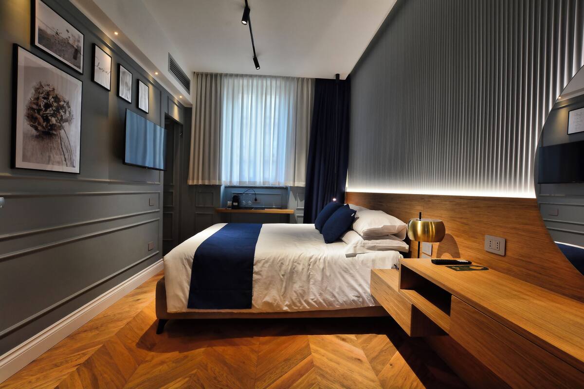 Una habitación de hotel moderna con una cama grande, cabecera de madera, mesitas de noche y un televisor de pantalla plana montado en la pared.