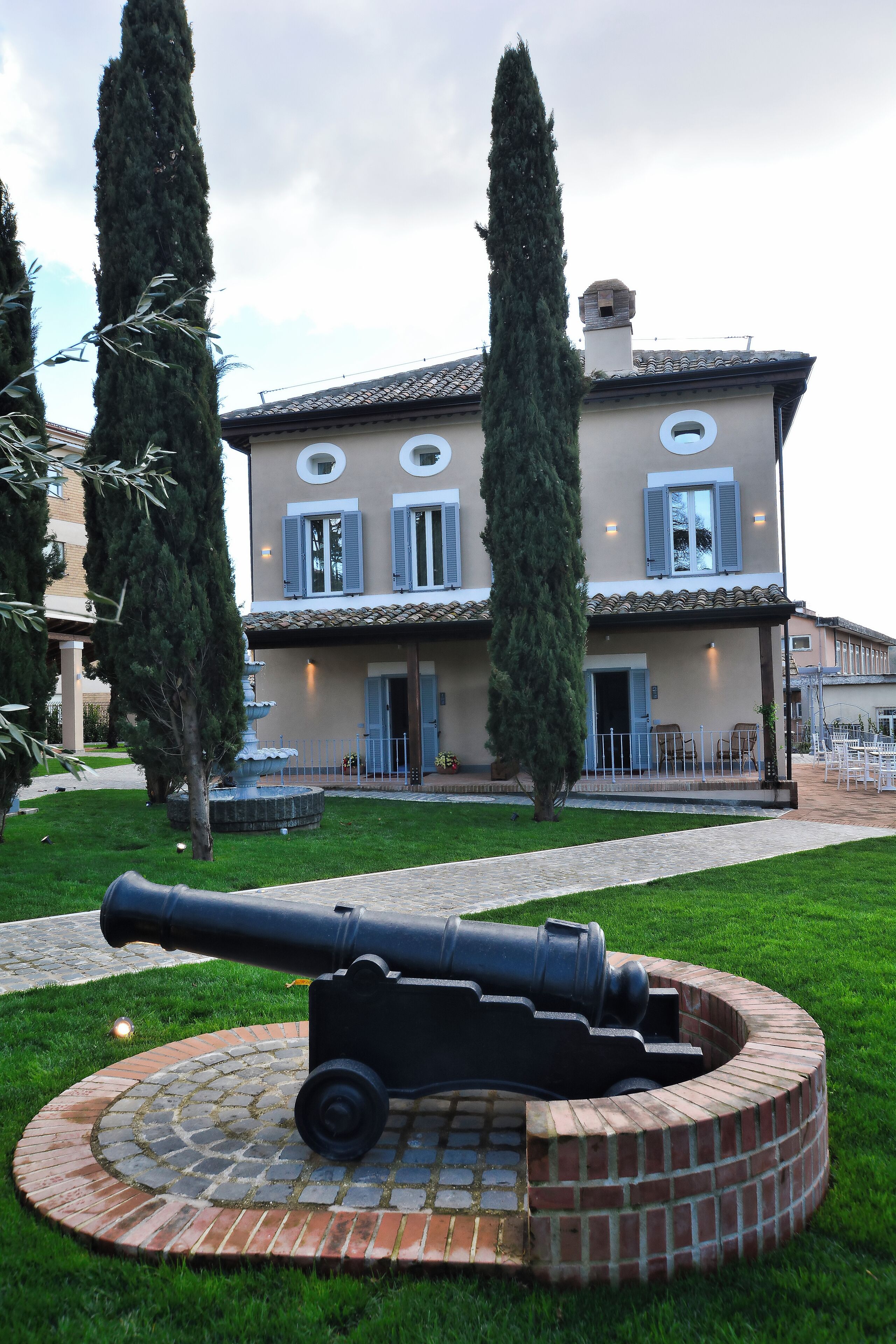 A building with a fountain, a cannon, and two tall trees in the foreground.